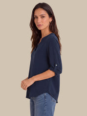 A woman wearing a navy blue V-neck top with rolled-up sleeves and blue jeans, standing against a solid beige background.
