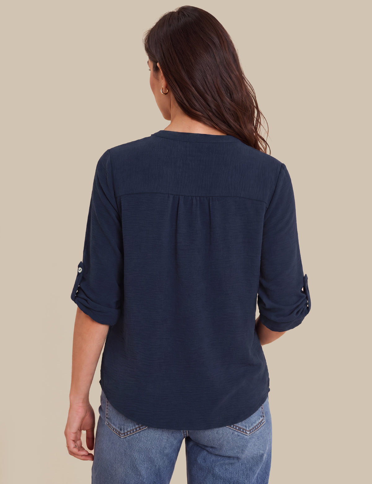 A woman viewed from the back wearing a navy blue three-quarter sleeve blouse with rolled cuffs and blue jeans, set against a solid beige background.