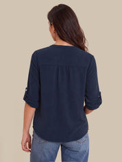 A woman viewed from the back wearing a navy blue three-quarter sleeve blouse with rolled cuffs and blue jeans, set against a solid beige background.