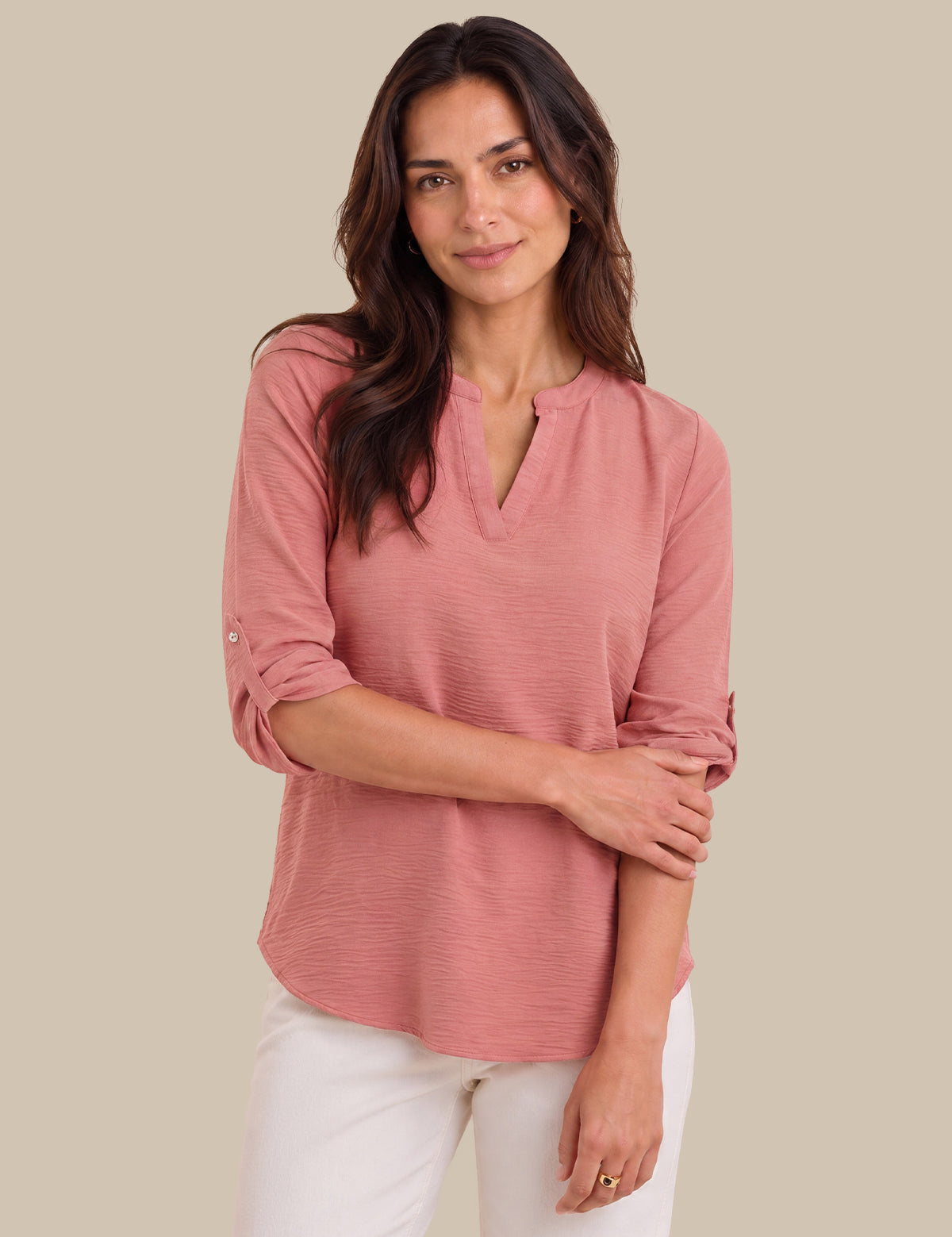 A woman modeling a dusty rose pink long-sleeve top with a split V-neckline and rolled sleeves, paired with white pants, against a solid beige background.