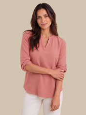 A woman modeling a dusty rose pink long-sleeve top with a split V-neckline and rolled sleeves, paired with white pants, against a solid beige background.