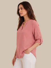 A woman wearing a dusty pink, three-quarter sleeve henley top with rolled-up sleeves and white pants, shown from a side profile against a solid beige background.