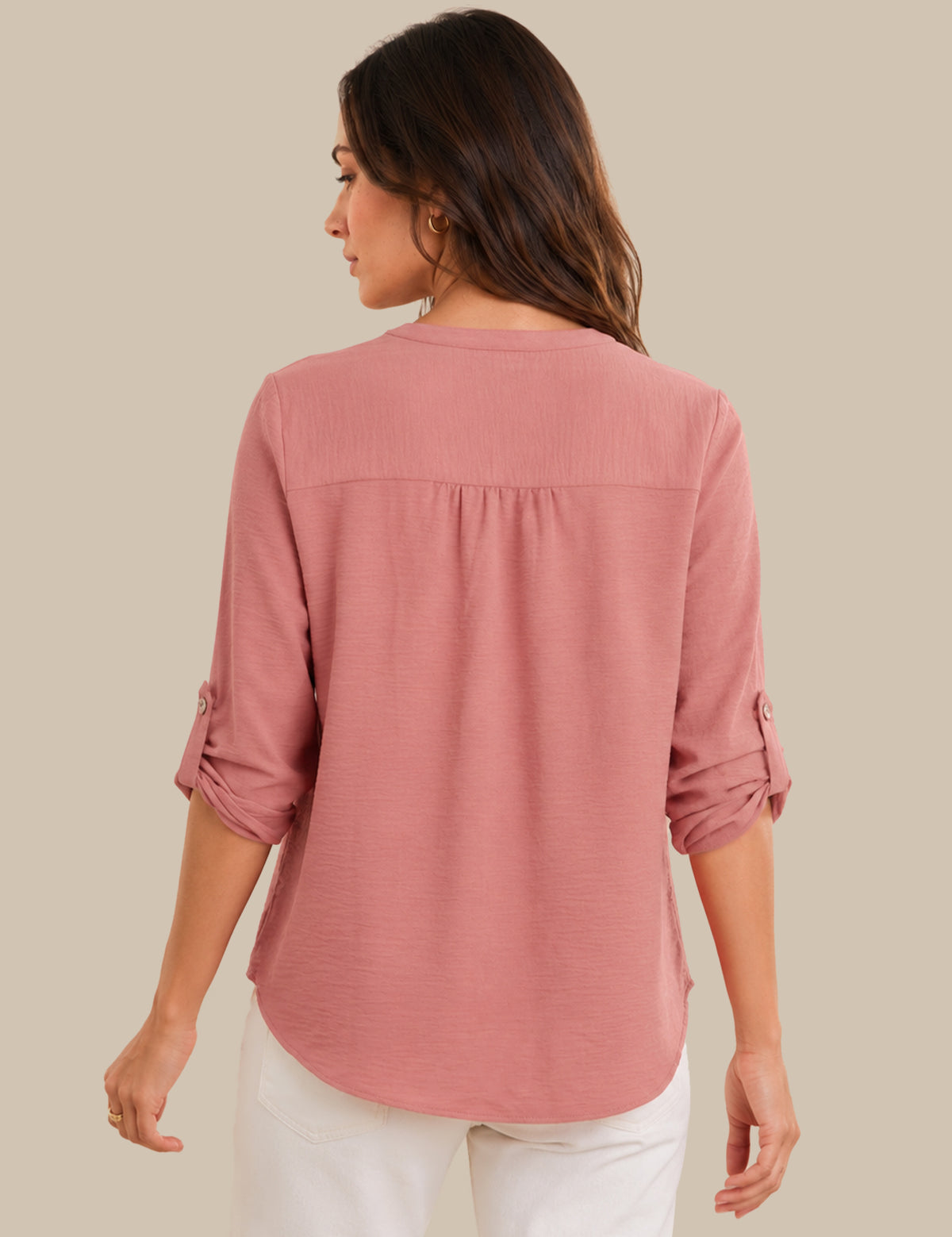 A woman shown from the back wearing a dusty rose pink long-sleeve blouse with rolled-up sleeves and white pants, set against a solid beige background.