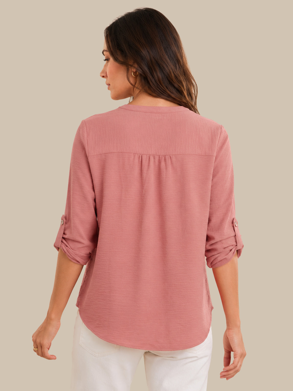 A woman shown from the back wearing a dusty rose pink long-sleeve blouse with rolled-up sleeves and white pants, set against a solid beige background.