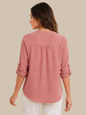 A woman shown from the back wearing a dusty rose pink long-sleeve blouse with rolled-up sleeves and white pants, set against a solid beige background.