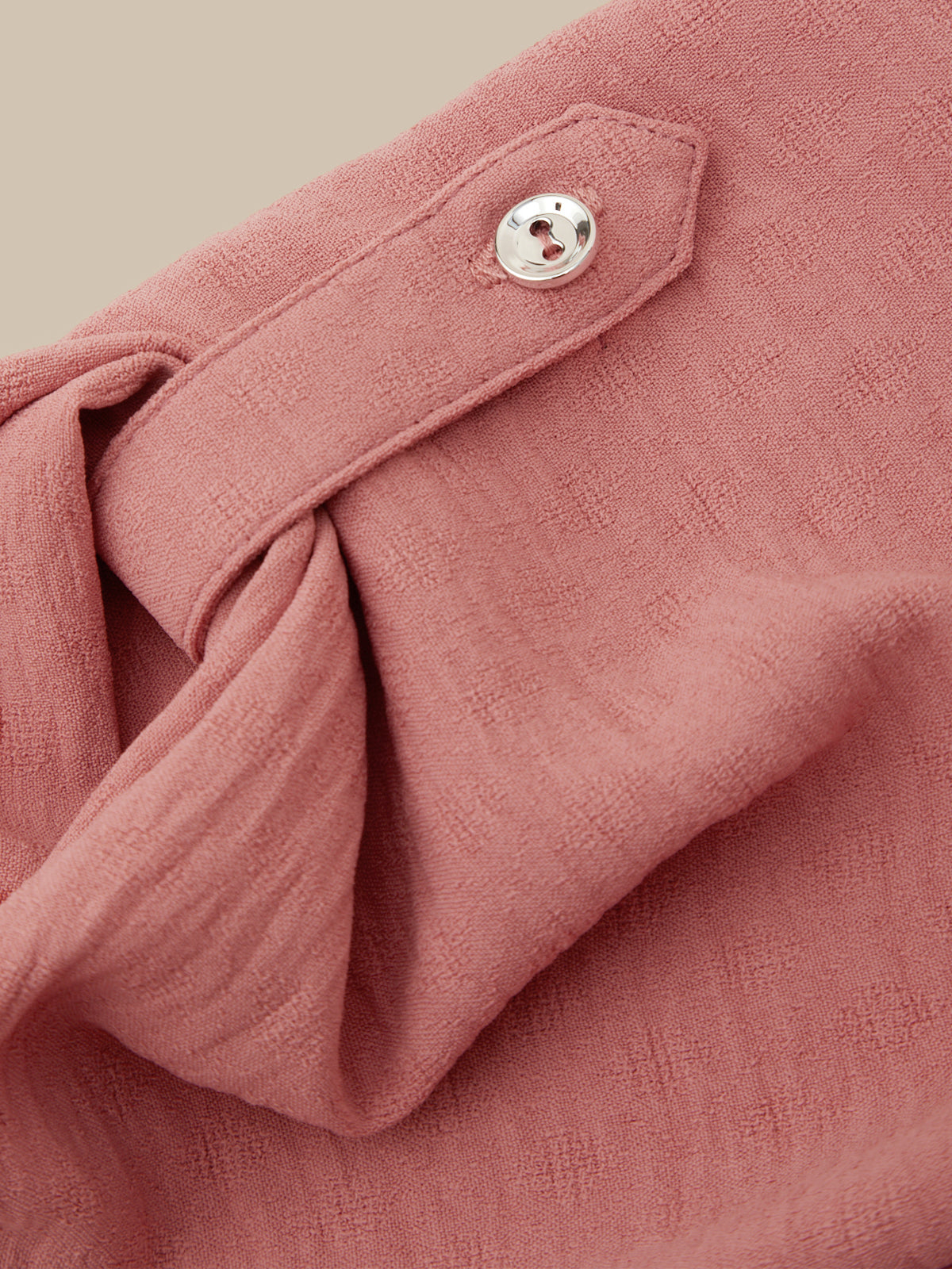 Close-up of a dusty rose pink textured fabric garment, showing a buttoned flap detail and soft folds against a warm beige background.
