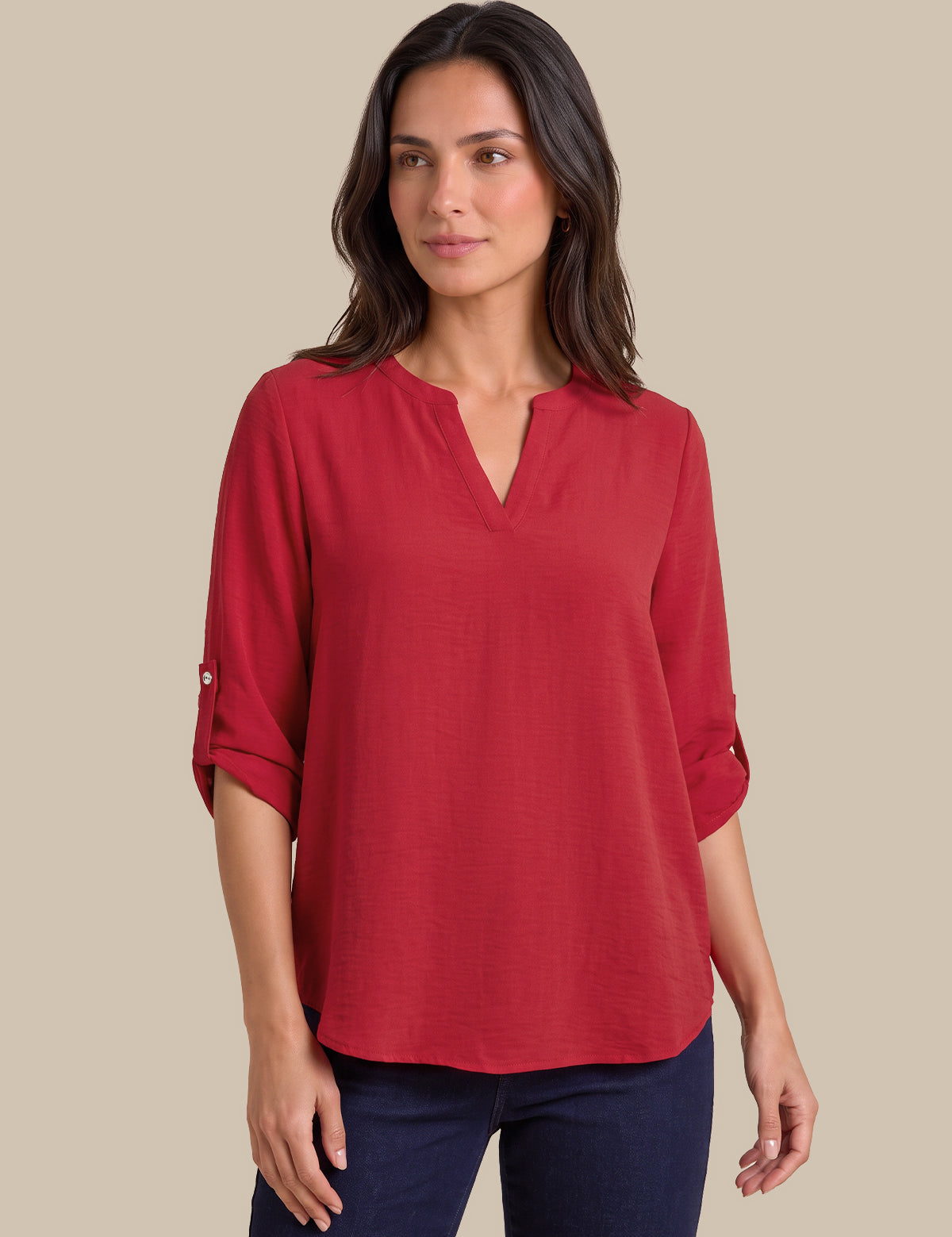 A woman wearing a red three-quarter sleeve blouse with a split V-neckline and rolled cuffs, paired with dark blue jeans, set against a solid beige background.