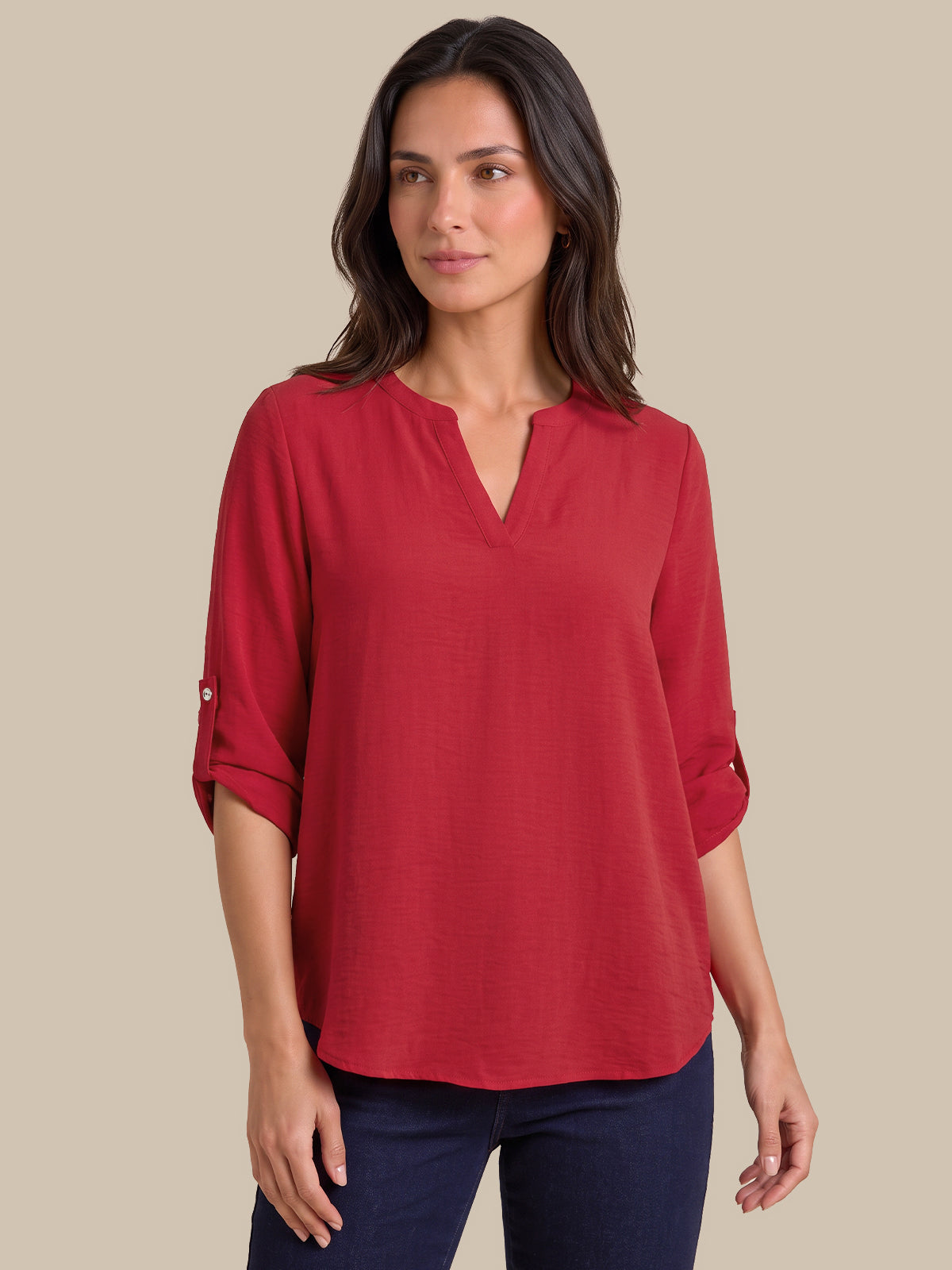 A woman wearing a red three-quarter sleeve blouse with a split V-neckline and rolled cuffs, paired with dark blue jeans, set against a solid beige background.