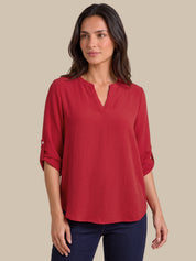 A woman wearing a red three-quarter sleeve blouse with a split V-neckline and rolled cuffs, paired with dark blue jeans, set against a solid beige background.