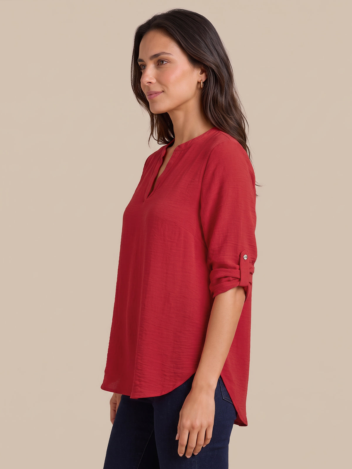A woman wearing a red, three-quarter sleeve top with rolled cuffs and a split neckline, paired with dark blue jeans, shown from a side profile against a solid beige background.