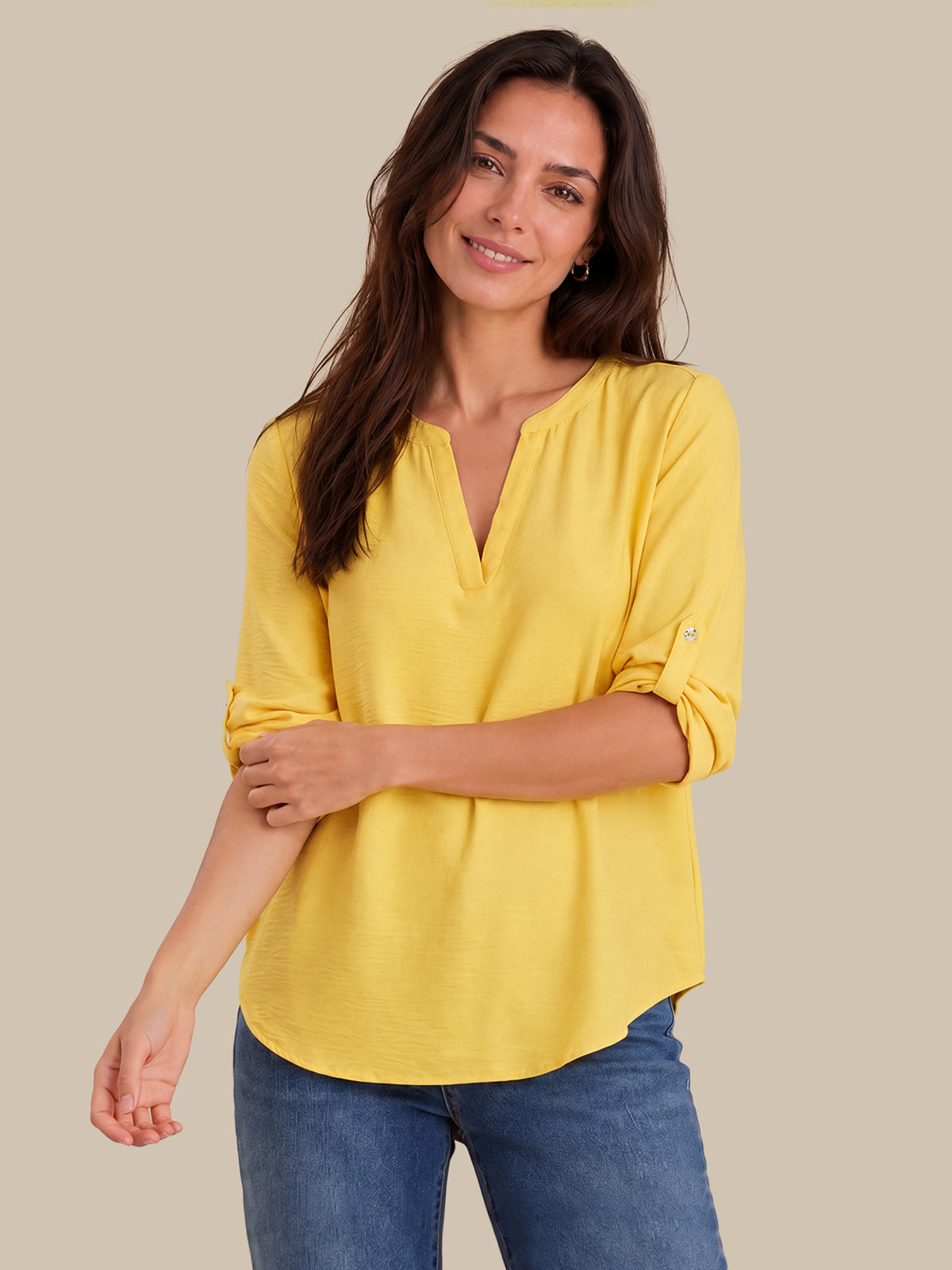 A woman wearing a bright yellow, three-quarter sleeve top with a split V-neckline and rolled-up sleeves, paired with blue jeans, set against a solid beige background.