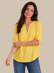 A woman wearing a bright yellow, three-quarter sleeve top with a split V-neckline and rolled-up sleeves, paired with blue jeans, set against a solid beige background.