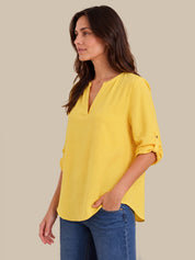 A woman wearing a bright yellow V-neck blouse with rolled sleeves and blue jeans, standing against a solid beige background.