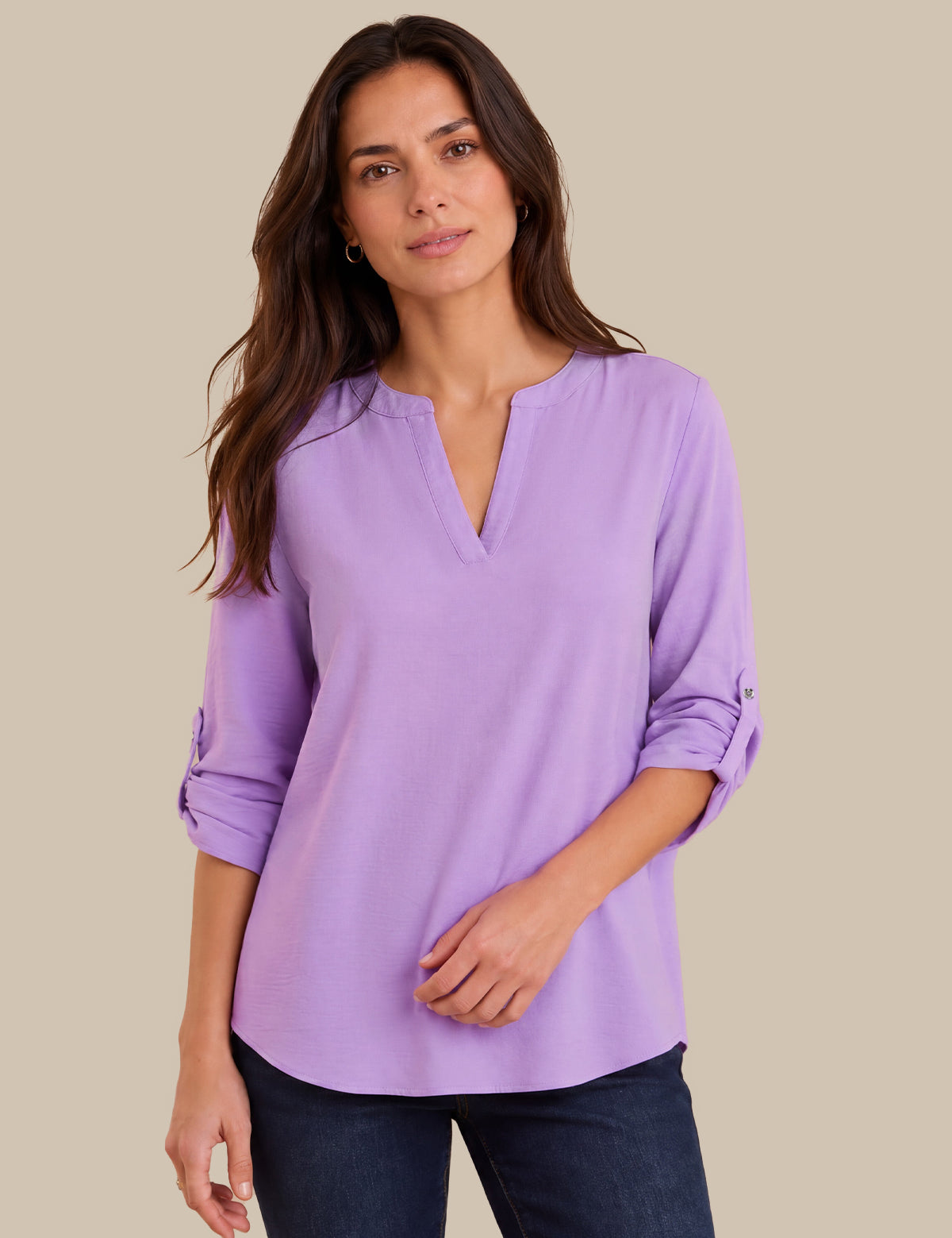 A woman modeling a lavender purple long-sleeve blouse with a split V-neckline and rolled sleeves, paired with dark blue jeans, against a solid beige background.