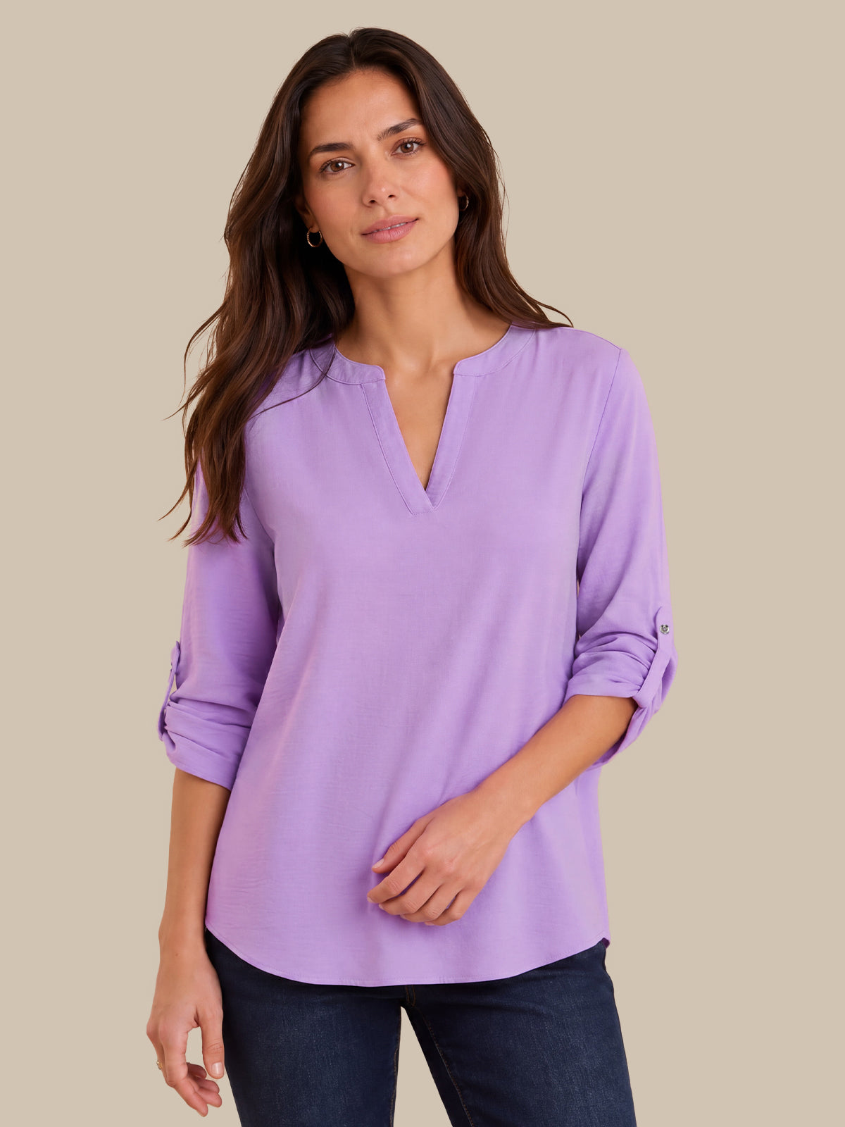 A woman modeling a lavender purple long-sleeve blouse with a split V-neckline and rolled sleeves, paired with dark blue jeans, against a solid beige background.