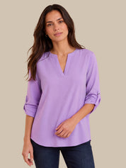 A woman modeling a lavender purple long-sleeve blouse with a split V-neckline and rolled sleeves, paired with dark blue jeans, against a solid beige background.