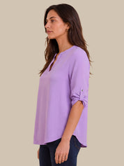 A woman shown in side profile wearing a lavender purple long-sleeve top with rolled-up sleeves and dark blue jeans, set against a solid beige background.