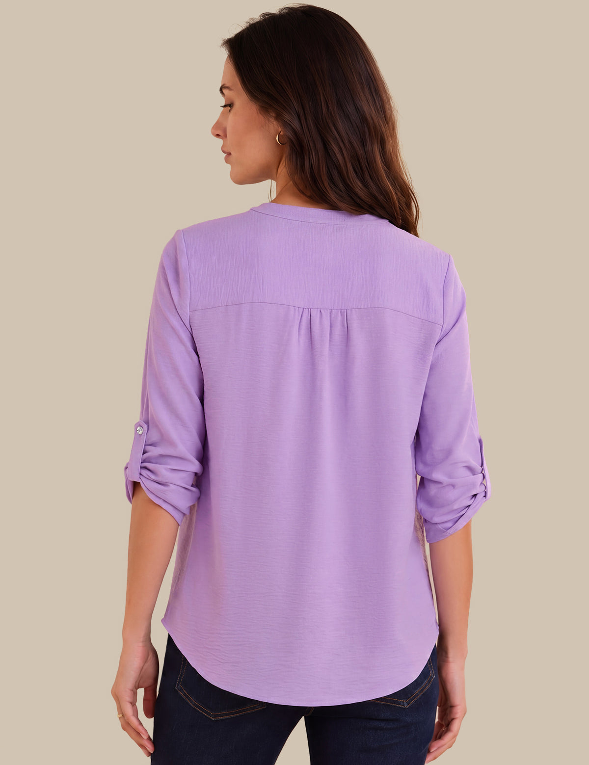 A woman shown from the back wearing a lavender purple long-sleeve blouse with rolled-up sleeves and dark blue jeans, set against a solid beige background.