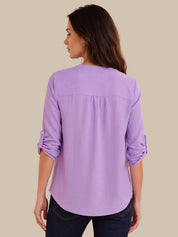 A woman shown from the back wearing a lavender purple long-sleeve blouse with rolled-up sleeves and dark blue jeans, set against a solid beige background.