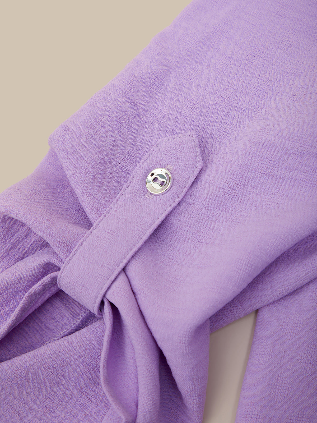 Close-up of a lavender purple garment with a buttoned flap detail, showing the textured fabric and a small silver button against a light beige background.