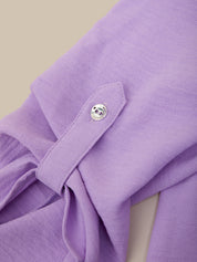 Close-up of a lavender purple garment with a buttoned flap detail, showing the textured fabric and a small silver button against a light beige background.