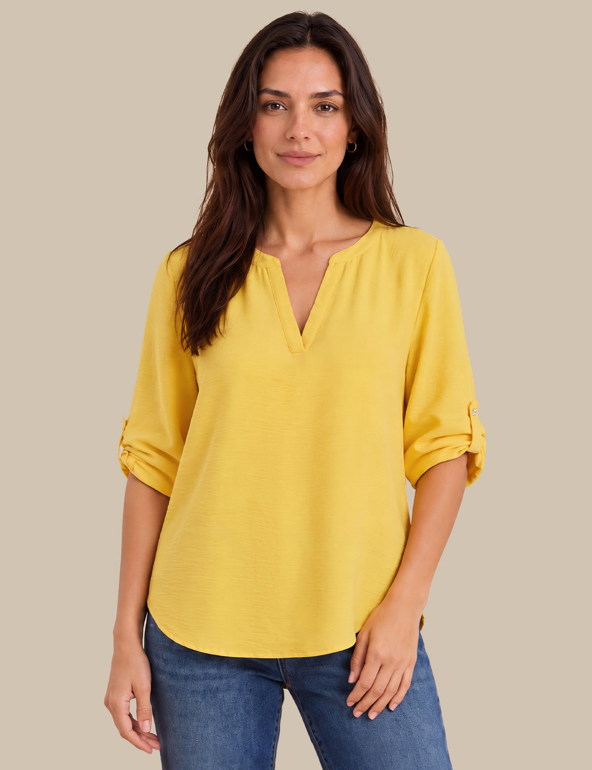 A woman wearing a bright yellow V-neck blouse with rolled three-quarter sleeves and blue jeans, standing against a solid beige background.