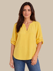 A woman wearing a bright yellow V-neck blouse with rolled three-quarter sleeves and blue jeans, standing against a solid beige background.
