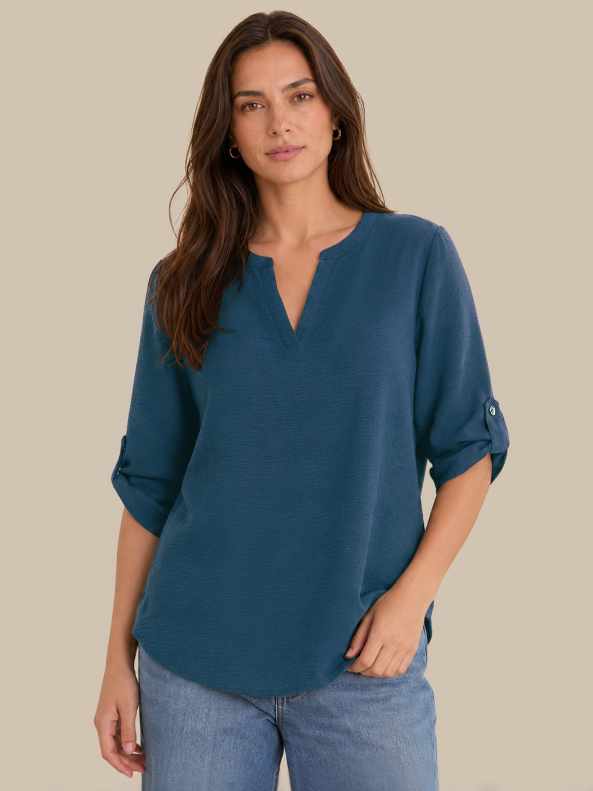 A woman modeling a slate blue V-neck blouse with rolled three-quarter sleeves, paired with light-wash blue jeans, against a solid beige background.