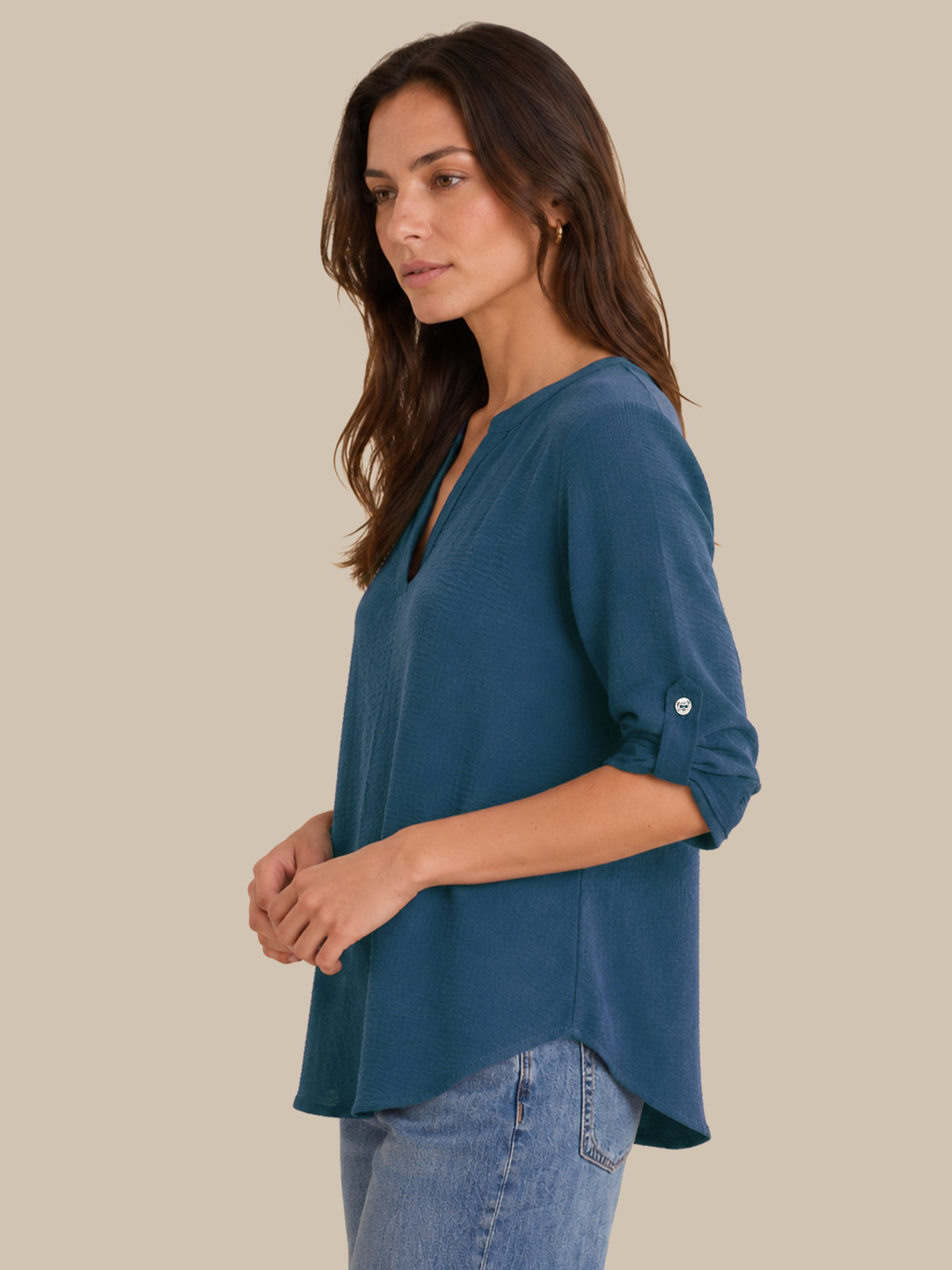 A woman wearing a slate blue V-neck top with rolled-up sleeves and light-wash blue jeans, shown from a side profile against a solid beige background.