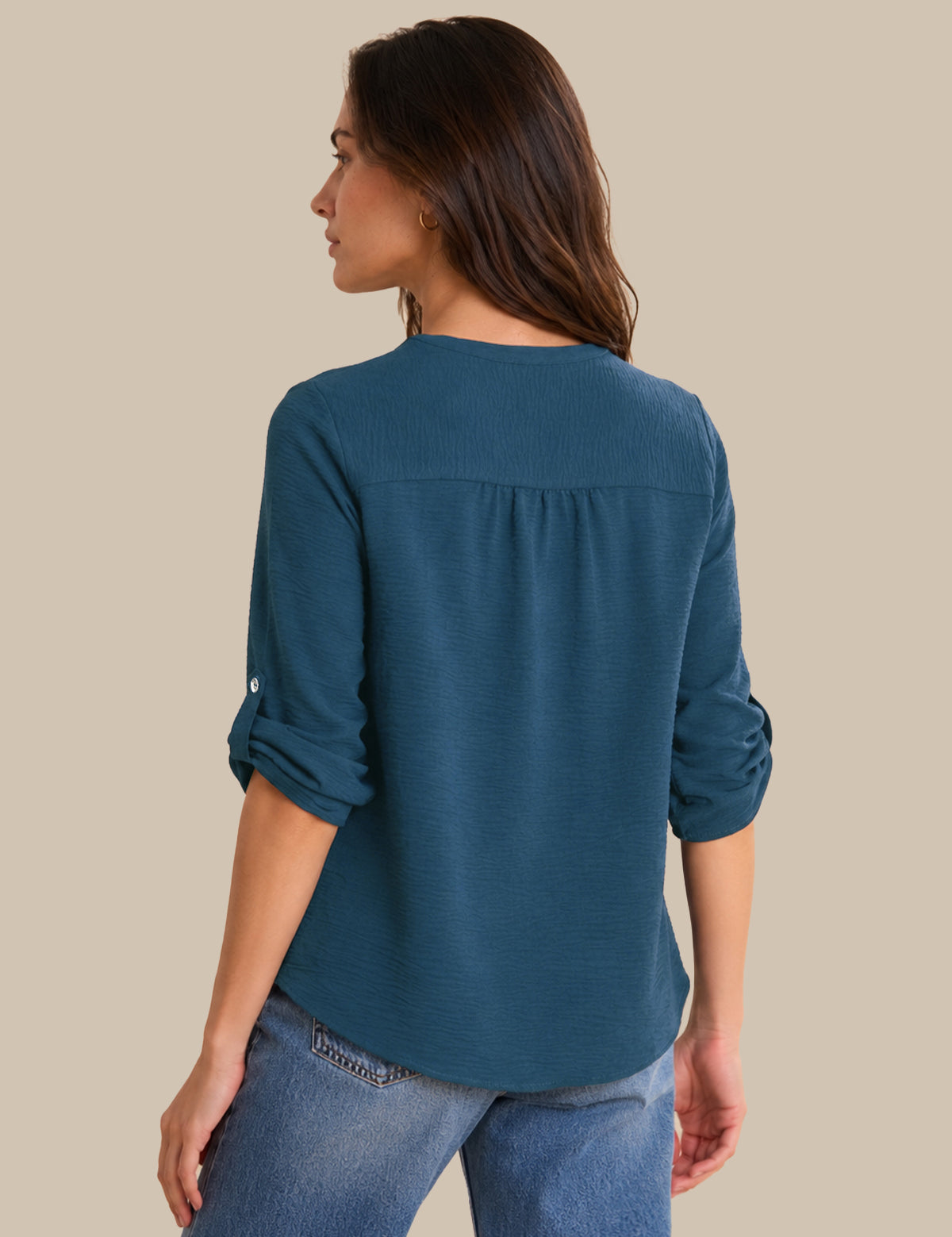A woman shown from the back wearing a teal blue long-sleeve top with rolled-up sleeves and blue jeans, set against a solid beige background.