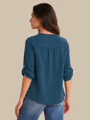 A woman shown from the back wearing a teal blue long-sleeve top with rolled-up sleeves and blue jeans, set against a solid beige background.