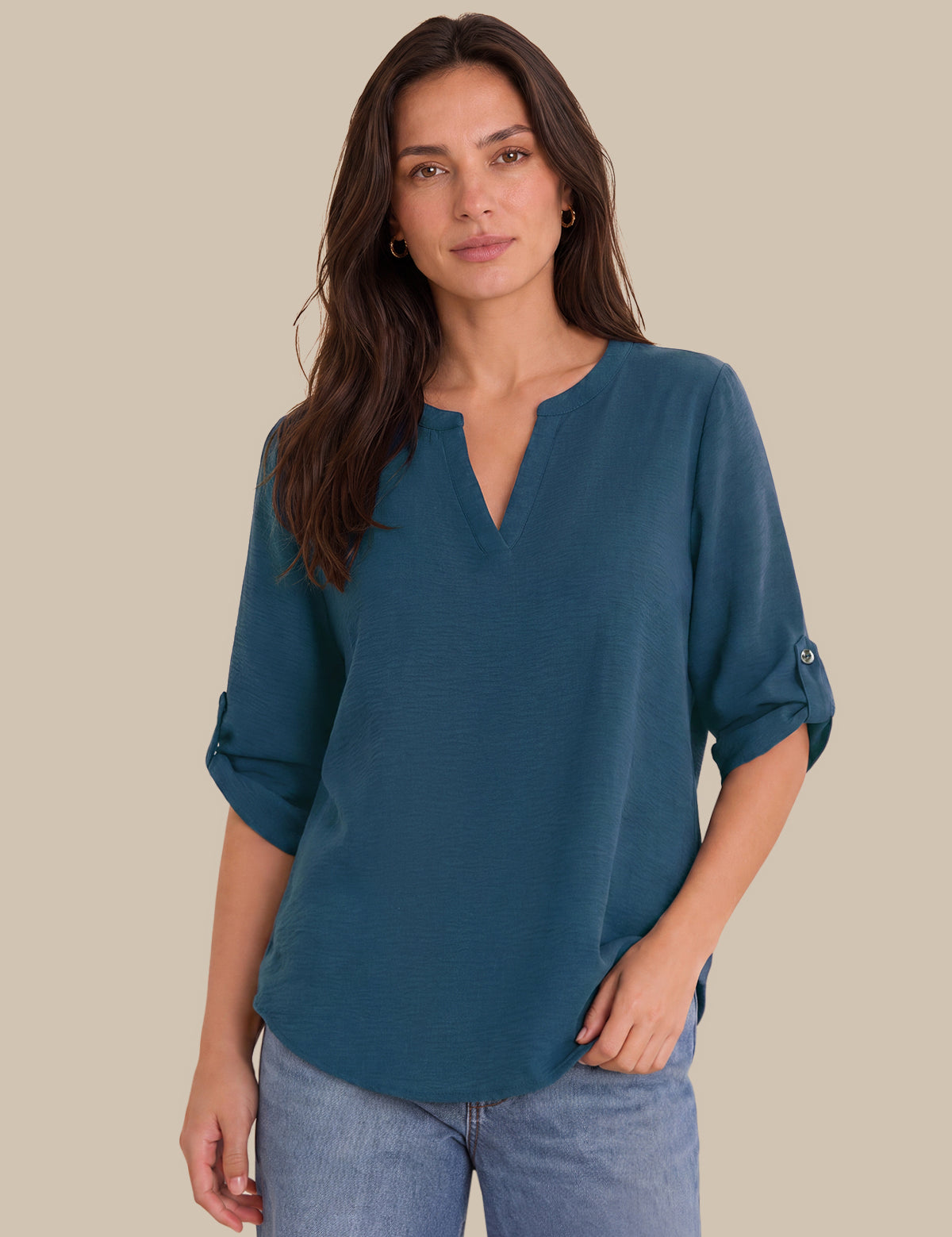 A woman wearing a teal blue V-neck blouse with rolled sleeves, paired with light blue jeans, standing against a solid beige background.