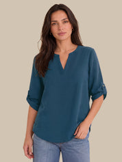 A woman wearing a teal blue V-neck blouse with rolled sleeves, paired with light blue jeans, standing against a solid beige background.