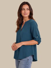 A woman modeling a teal blue V-neck blouse with three-quarter sleeves and rolled cuffs, paired with light-wash blue jeans, against a solid beige background.