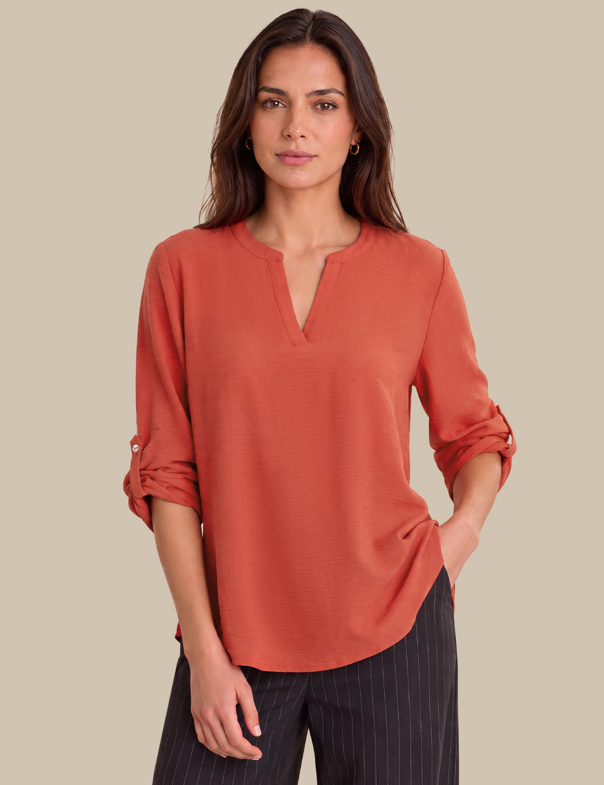 A woman modeling a rust-orange long-sleeve blouse with a split V-neck and rolled sleeves, paired with dark pinstriped trousers, against a solid beige background.