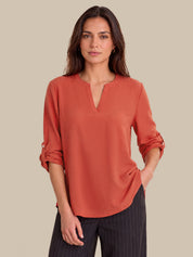 A woman modeling a rust-orange long-sleeve blouse with a split V-neck and rolled sleeves, paired with dark pinstriped trousers, against a solid beige background.