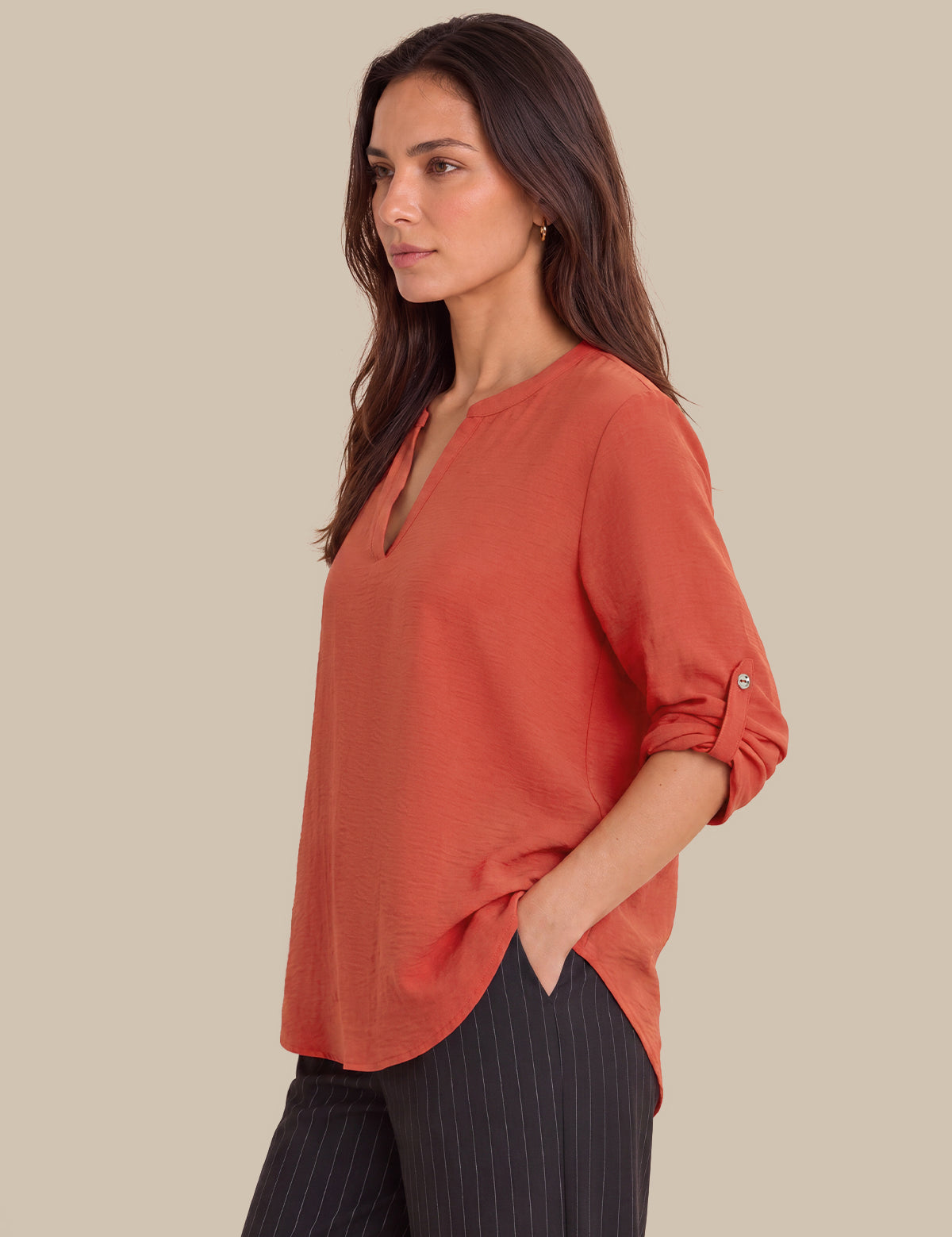 A woman wearing a rust-orange long-sleeve blouse with rolled cuffs and dark pinstripe trousers, photographed against a solid beige studio background.