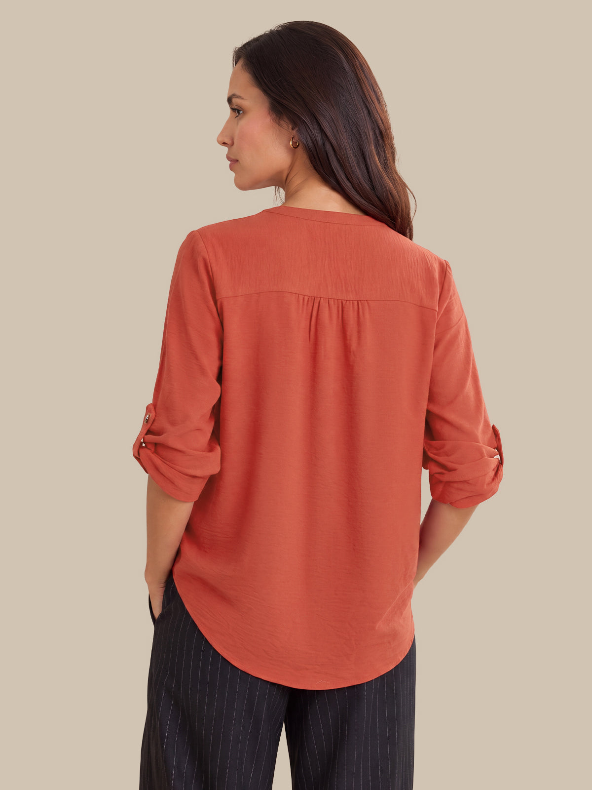 A woman shown from the back wearing a rust-orange long-sleeve blouse with rolled cuffs and dark pinstriped trousers, set against a solid beige background.