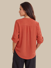 A woman shown from the back wearing a rust-orange long-sleeve blouse with rolled cuffs and dark pinstriped trousers, set against a solid beige background.