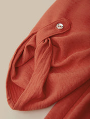 Close-up of a terracotta rust-colored textured fabric garment with a small silver button detail, set against a solid beige background.