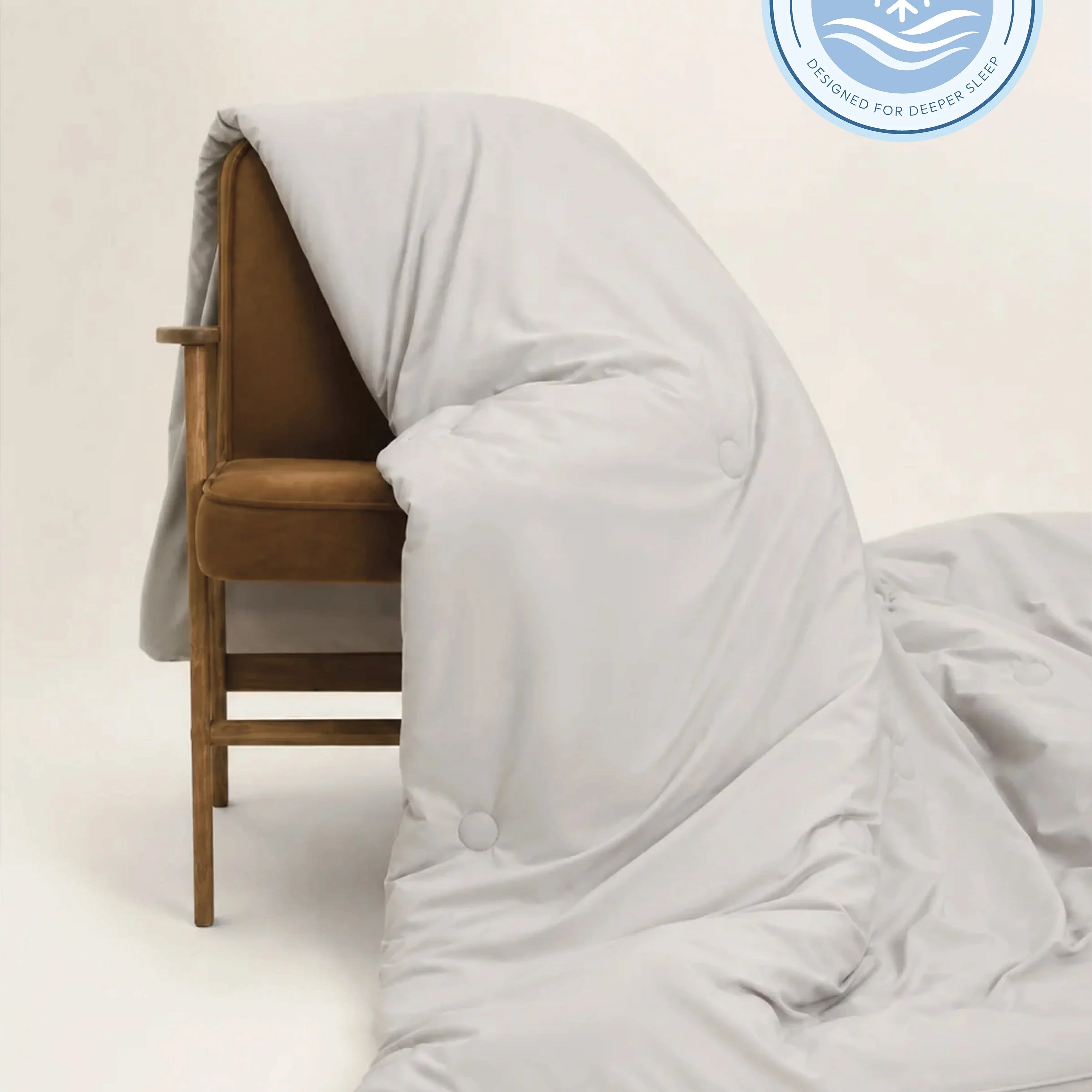 A light gray cooling duvet or comforter draped over a brown upholstered wooden chair, with a circular cooling technology badge in the top right corner.
