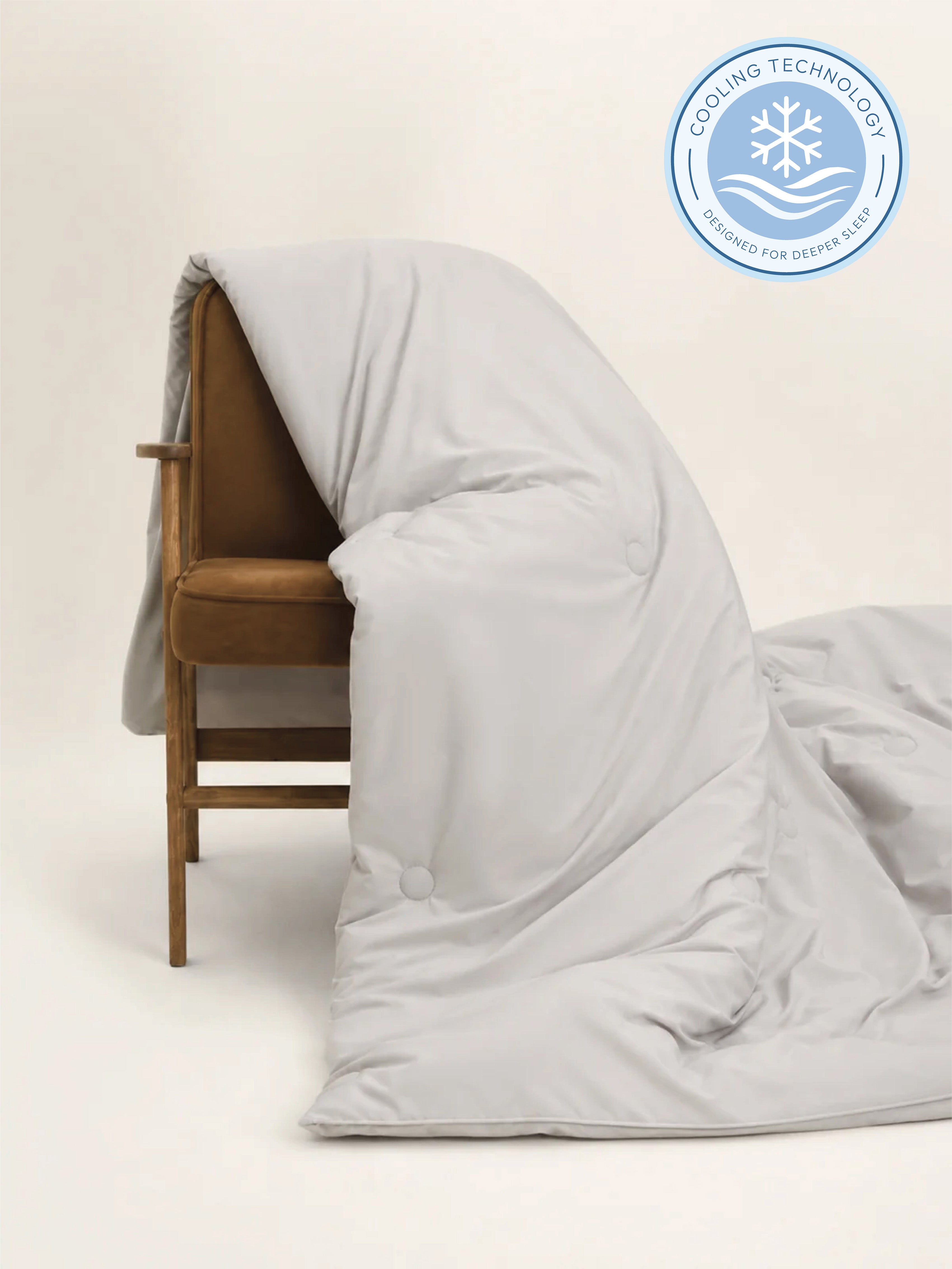 A light gray cooling duvet or comforter draped over a brown upholstered wooden chair, with a circular cooling technology badge in the top right corner.