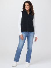 Woman wearing a black vest, white long-sleeve shirt, blue jeans, and white sneakers on a white background