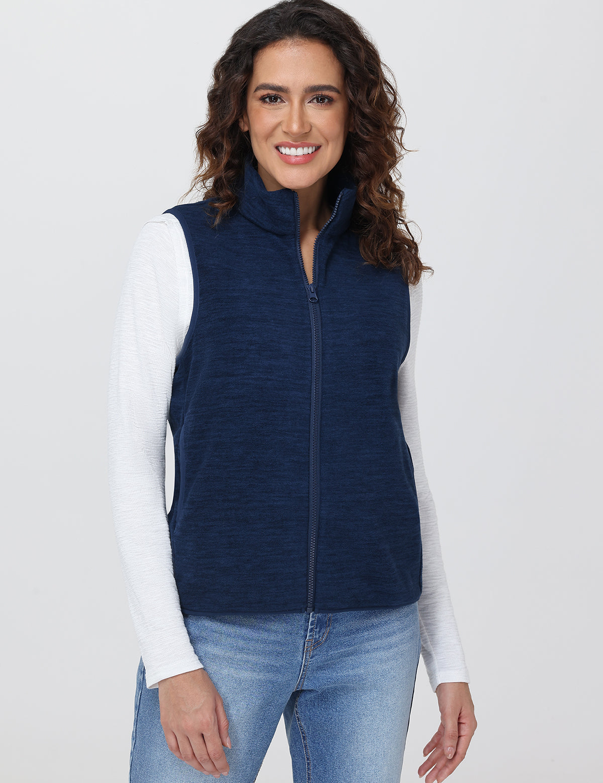 Woman wearing a navy blue vest over a white long-sleeve shirt and blue jeans on a light gray background