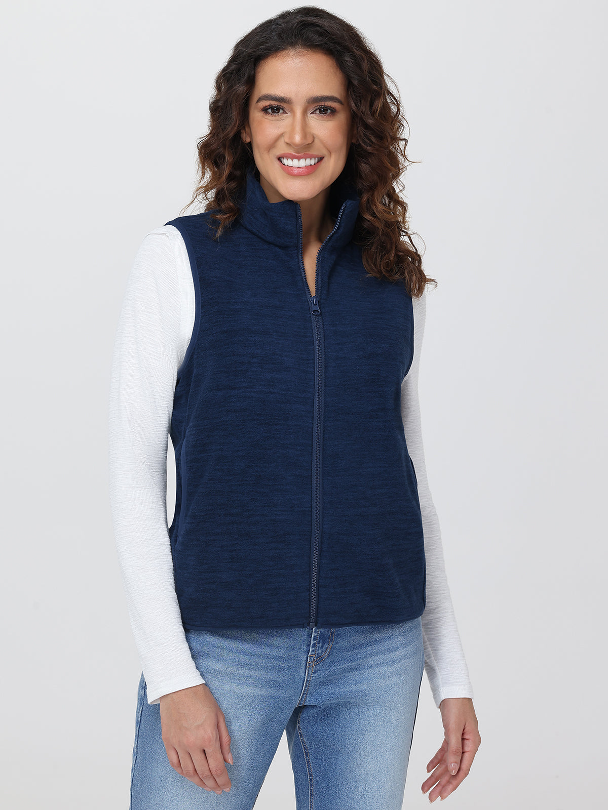 Woman wearing a navy blue vest over a white long-sleeve shirt and blue jeans on a light gray background