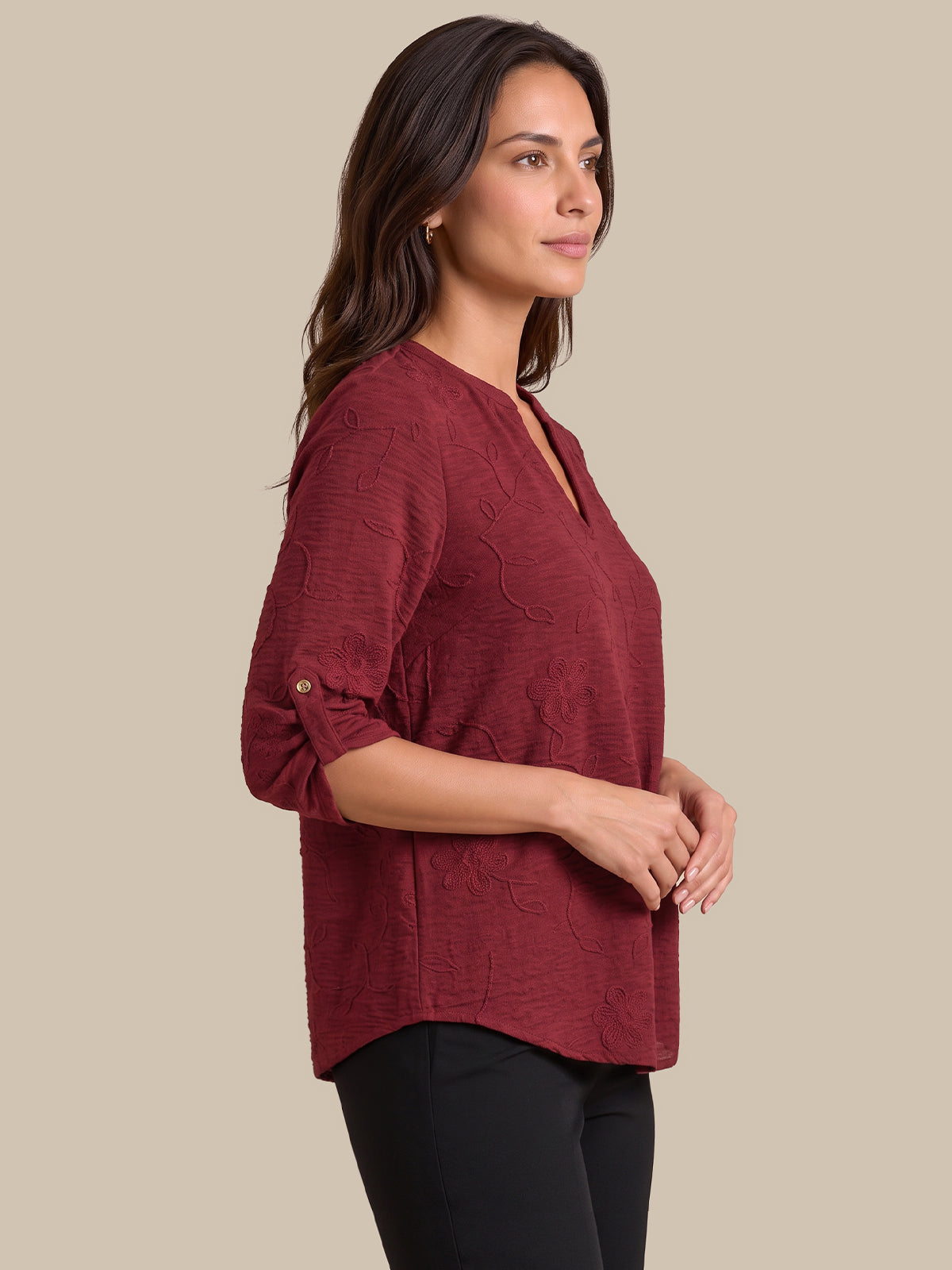 A woman wearing a textured, deep burgundy V-neck top with three-quarter sleeves and black pants, shown in profile against a solid beige background.