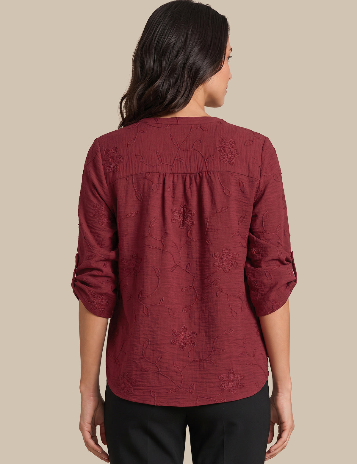 A woman viewed from the back wearing a textured, crinkled burgundy long-sleeve blouse with a gathered neckline, paired with black pants, against a solid beige background.
