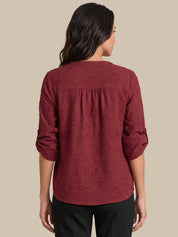 A woman viewed from the back wearing a textured, crinkled burgundy long-sleeve blouse with a gathered neckline, paired with black pants, against a solid beige background.