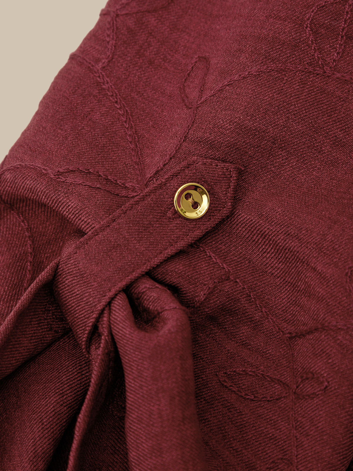 Close-up of a burgundy textured fabric garment featuring a small gold button closure and tonal embroidered leaf patterns.
