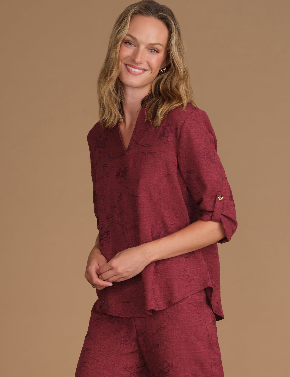 A woman modeling a matching burgundy tops with a subtle tonal floral jacquard pattern, featuring a short-sleeve top with rolled cuffs and matching pants, set against a solid tan studio background.